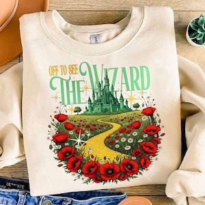 Op de afbeelding: Crèmekleurige sweatshirt met een kleurrijke afbeelding van een gele bakstenen weg die naar een groen kasteel leidt. De tekst "Off to See the Wizard" is in groen en geel. Rode klaprozen en witte bloemen omringen de weg.