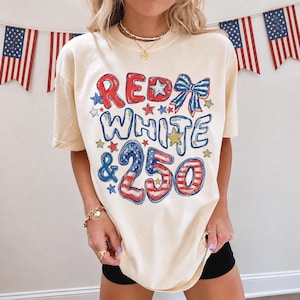 Op de afbeelding: Crèmekleurig T-shirt met de tekst "RED WHITE & 250" in rood, wit en blauw. Het ontwerp bevat sterren en een strik, ter viering van 4 juli. Amerikaanse vlaggen op de achtergrond.
