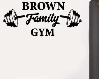 May include: A white wall features the black text "BROWN Family GYM" with barbell illustrations. A black punching bag hangs from the ceiling. Dumbbells are on the floor, indicating a home gym. The image conveys a fitness and family theme.