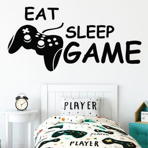 Op de afbeelding: Een witte slaapkamer met een bed met een dekbedovertrek en kussen met een videogamecontroller-thema. Op de muur boven het bed staan de woorden "EAT SLEEP GAME" in het zwart. Een kleine bijzettafel heeft een klok.