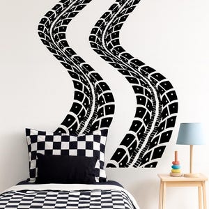 May include: Black tire track wall decals in a wavy pattern. A black and white checkered pillow and bedspread are visible, along with a bedsheet featuring cartoon race cars. A small wooden side table with a lamp and a stack of colorful rings is also present.