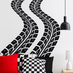May include: Black and white tire track wall decals on a white wall above a bed with red, black, and checkered pillows. A black pendant light and a white bedside lamp are also visible.