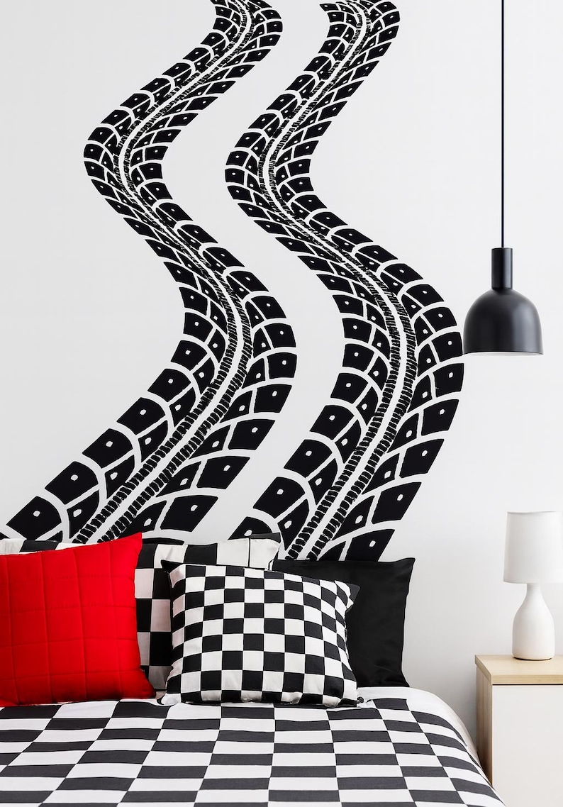 May include: Black and white tire track wall decals on a white wall above a bed with red, black, and checkered pillows. A black pendant light and a white bedside lamp are also visible.