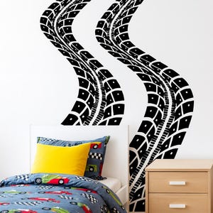 May include: Black tire track wall decals in a bedroom setting. The decals curve across a white wall, above a bed with a blue race car themed comforter and a yellow pillow. A wooden nightstand is to the right.