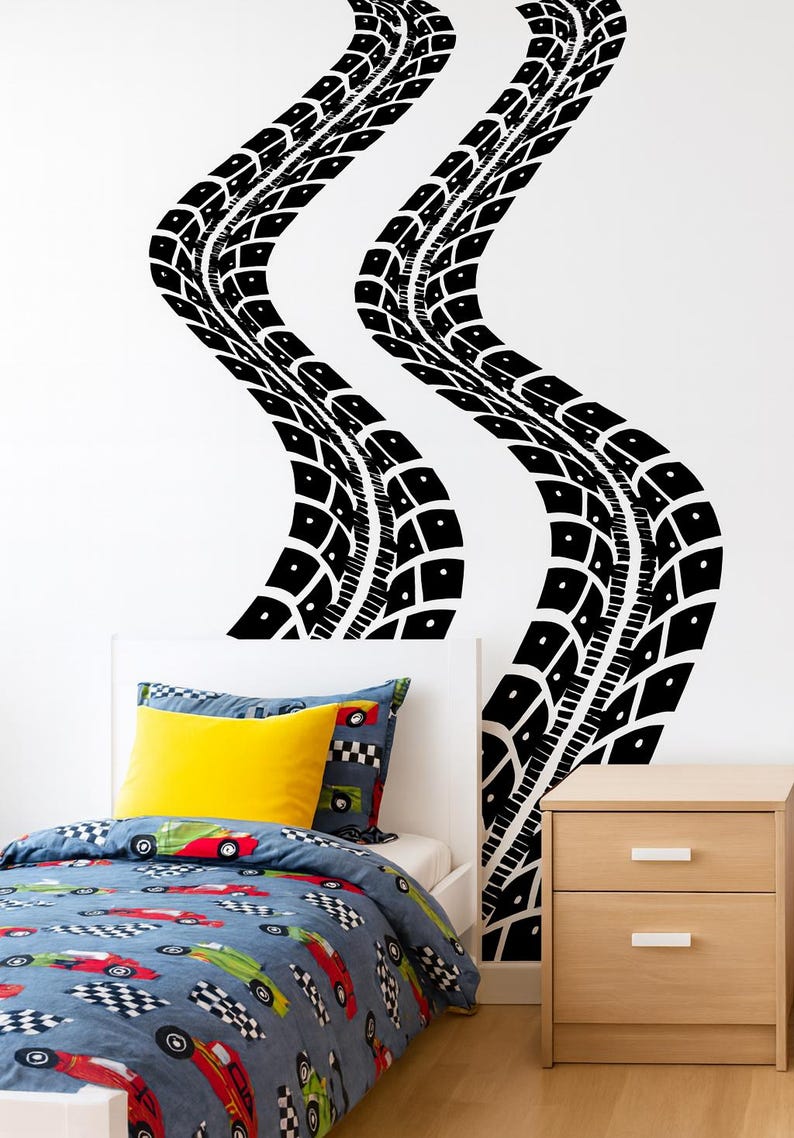 May include: Black tire track wall decals in a bedroom setting. The decals curve across a white wall, above a bed with a blue race car themed comforter and a yellow pillow. A wooden nightstand is to the right.