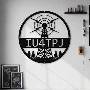 Peut inclure: Décoration murale en métal noir représentant une tour radio dans un cercle. Le texte "IU4TPJ" est affiché. Le design comprend des éclairs, des cercles concentriques et des arbres stylisés, idéal pour une maison ou un bureau.