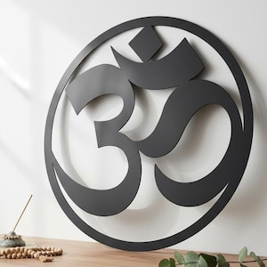 May include: A black metal Om symbol wall art. The symbol is enclosed in a circular frame. The Om symbol is a spiritual icon in Hinduism, Buddhism, and Jainism. The wall art is mounted on a white wall.