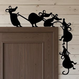 Peut inclure: Silhouette noire en métal d'un groupe de souris grimpant et suspendues à un cadre de porte. Les souris sont représentées dans diverses poses, certaines tirant une corde et d'autres suspendues à l'envers. La porte est marron et le mur est en bois clair.