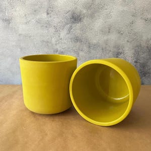 May include: Two yellow ceramic planters. One is upright, the other is on its side, revealing the interior. The planters have a smooth, matte finish and a rounded shape. They are set against a neutral background.