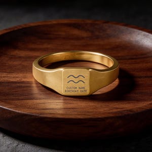Puede incluir: Un anillo de sello dorado con una cara cuadrada con dos líneas onduladas y el texto "CUSTOM NAME BIRTHDAY DATE". El anillo se muestra en un plato de madera oscura.