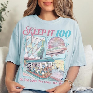 May include: Light blue t-shirt with a graphic featuring Mickey Mouse, Minnie Mouse, Donald Duck, and other characters on a boat. The shirt has the text "KEEP IT 100" and "On The Land, The Seas, The Sky".