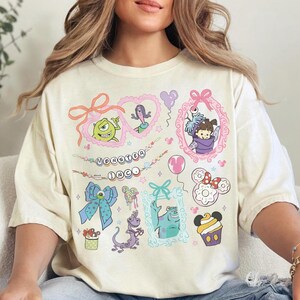 Può includere: T-shirt color avorio con grafica di personaggi dei cartoni animati colorati. La maglietta ha girocollo e maniche corte. La grafica include personaggi di "Monsters & Co." e altri personaggi Disney. Il testo "Monster Inc." è anch'esso visibile.