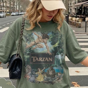 Op de afbeelding: Mosgroen T-shirt met een Tarzan-afbeelding. Het ontwerp bevat het woord "Tarzan" in een rechthoekige lijst, met illustraties van Tarzan en andere personages. Het shirt heeft een relaxte pasvorm.