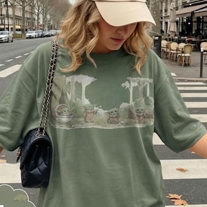Puede incluir: Camiseta verde musgo con un estampado de escena de bosque con varios personajes de Grogu. La camiseta tiene mangas cortas y un corte holgado. La palabra "Moss" est&aacute; escrita en cursiva en la esquina inferior izquierda.
