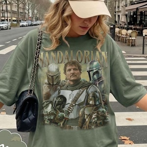 Pu&ograve; includere: Una t-shirt verde oliva con una stampa grafica dei personaggi di The Mandalorian, tra cui una figura centrale con i baffi, varie figure corazzate con elmo e una piccola creatura verde. &Egrave; visibile anche una borsa a tracolla nera trapuntata.