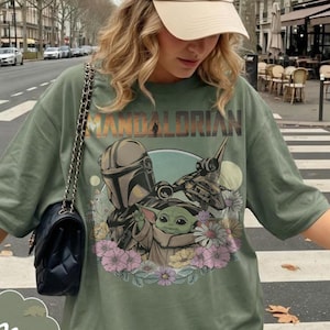 May include: Olive green t-shirt featuring a graphic of The Mandalorian and Grogu surrounded by flowers. The text "MANDALORIAN" is printed above the image. The shirt is a relaxed fit.