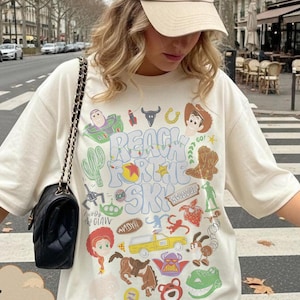 Pode incluir: Camiseta cor marfim com um design gr&aacute;fico colorido com personagens de desenhos animados e o texto "REACH FOR THE SKY". A camiseta &eacute; combinada com um bon&eacute; de beisebol bege e uma bolsa preta com al&ccedil;a de corrente. A camiseta tem um ajuste folgado.