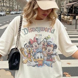 Pu&ograve; includere: T-shirt color avorio con grafica Disneyworld con Topolino, Minnie, Paperino e altri personaggi. Il testo "Disneyworld 2026" &egrave; stampato sopra i personaggi. La maglietta &egrave; abbinata a un berretto da baseball beige e una borsa nera.