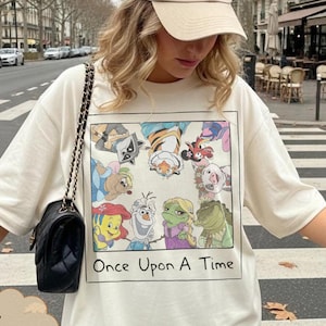 May include: Ivory-colored t-shirt featuring a colorful cartoon graphic with the text "Once Upon A Time." The graphic includes various animated characters. The shirt is paired with a beige baseball cap and a black purse with a chain strap.