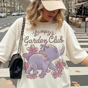 Op de afbeelding: Ivoor t-shirt met een paarse olifant afbeelding omringd door roze bloemen en de tekst "Jumpy's Garden Club". Het shirt wordt gedragen met een beige baseball cap.