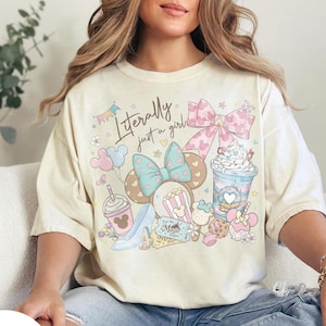 May include: Ivory-coloured t-shirt with a whimsical graphic featuring a bow, Mickey Mouse ears, a high heel, popcorn, and a drink. The text "Literally just a girl" is printed on the front. The shirt is a relaxed fit.