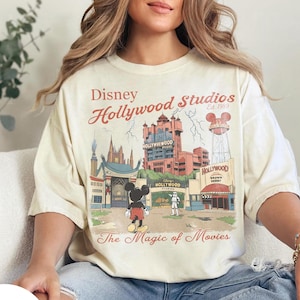 Può includere: T-shirt color avorio con grafica Disney Hollywood Studios. Il design include illustrazioni di Mickey Mouse, edifici iconici del parco e il testo "Disney Hollywood Studios Est. 1989" e "The Magic of Movies".