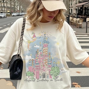 Puede incluir: Camiseta de color marfil con un gr&aacute;fico de castillo colorido y el texto "Happiest Birthday on Earth". El dise&ntilde;o incluye un hada y velas. La camiseta se combina con una gorra de b&eacute;isbol beige y un bolso negro.