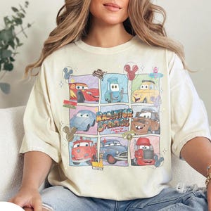 May include: Ivory-colored t-shirt featuring a grid of cartoon cars with the text "Radiator Springs". The design includes various car characters in colorful squares. The shirt is a relaxed fit.