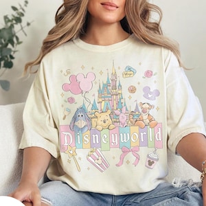 May include: Ivory-colored t-shirt featuring a colorful Disneyworld graphic. The design includes a castle, balloons, and various Disney characters like Winnie the Pooh and Tigger. The word "Disneyworld" is prominently displayed.