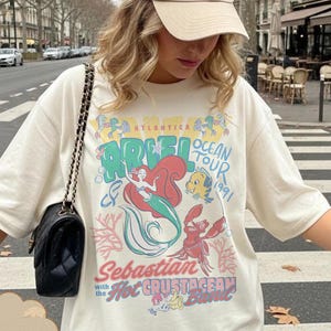 May include: Ivory-colored t-shirt featuring a graphic of Ariel from The Little Mermaid, with the text "Ariel Ocean Tour 1991." The design includes illustrations of Ariel, Flounder, and Sebastian. The shirt has short sleeves.