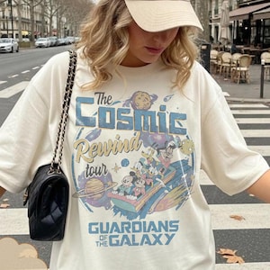 May include: Ivory-colored t-shirt featuring a graphic design with the text "The Cosmic Rewind Tour" and "Guardians of the Galaxy." The design includes cartoon characters and space-themed elements. The shirt has short sleeves and a relaxed fit.