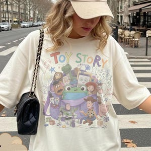 May include: An ivory short-sleeved t-shirt featuring a colourful graphic print of Toy Story characters including Woody, Buzz Lightyear, and a green frog. The text Toy Story is printed in multicoloured letters above the characters on the front.
