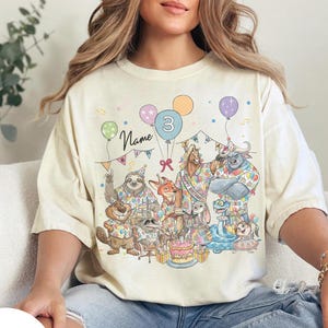 以下が含まれることがあります： アイボリー色のTシャツ。漫画の動物、風船、ケーキが描かれた、カラフルな誕生日のパーティーシーンが特徴です。「Name」と数字の「3」の文字も表示されています。このシャツは快適でカジュアルなスタイルです。