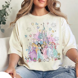 May include: Ivory-colored t-shirt featuring a colorful illustration of Disney princesses in front of a castle. The design includes fireworks, balloons, and floral accents. The word "IVORY" is printed on a white circle.