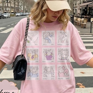 May include: A light pink t-shirt featuring a grid of cartoon illustrations of Disney princesses with their names and years. The shirt has short sleeves and a relaxed fit. The word "Blossom" is printed on the bottom left.