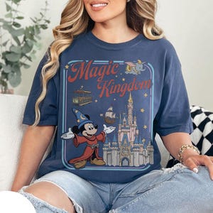 May include: Navy blue t-shirt with a retro graphic of Mickey Mouse as a sorcerer, and the words "Magic Kingdom" above a castle illustration. The shirt has a relaxed fit and is made of a soft material.