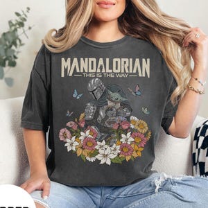 Puede incluir: Camiseta gris oscuro con el texto "MANDALORIAN - THIS IS THE WAY". El dise&ntilde;o muestra al Mandaloriano y a Grogu rodeados de flores y mariposas. Una prenda c&oacute;moda e informal.