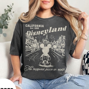 Może przedstawiać: Ciemnoszara koszulka z grafiką Disneyland. Grafika przedstawia Myszka Miki przed sceną uliczną z napisem "California Disneyland Est. 1955" i "The happiest place on earth."