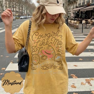 May include: A mustard yellow t-shirt featuring a graphic print with the text Cowgirl Like Me, a pink cowboy hat, a lasso, and star motifs. The shirt has short sleeves and a crew neckline, paired with a black quilted shoulder bag with a chain strap.