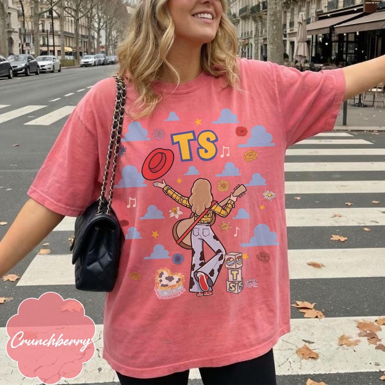 May include: A coral-colored short-sleeve t-shirt featuring a graphic print of a person with long hair playing a guitar, wearing a yellow plaid shirt and cow-print pants. The design includes blue clouds, yellow stars, a red hat, and the text TS.