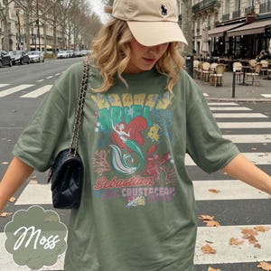 May include: Moss green t-shirt featuring a colorful graphic of a mermaid, fish, and lobster with the text "Ariel" and "Sebastian Crustacean". The shirt has short sleeves and a relaxed fit.