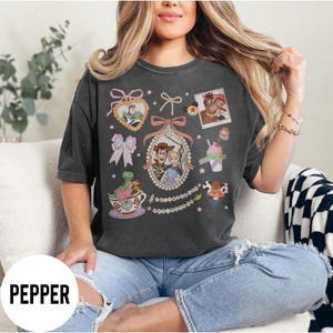 May include: Dark gray t-shirt featuring a collection of cartoon character illustrations. The design includes images of Woody, Buzz Lightyear, and other characters, along with decorative bows and a teacup. The word "PEPPER" is printed at the bottom.
