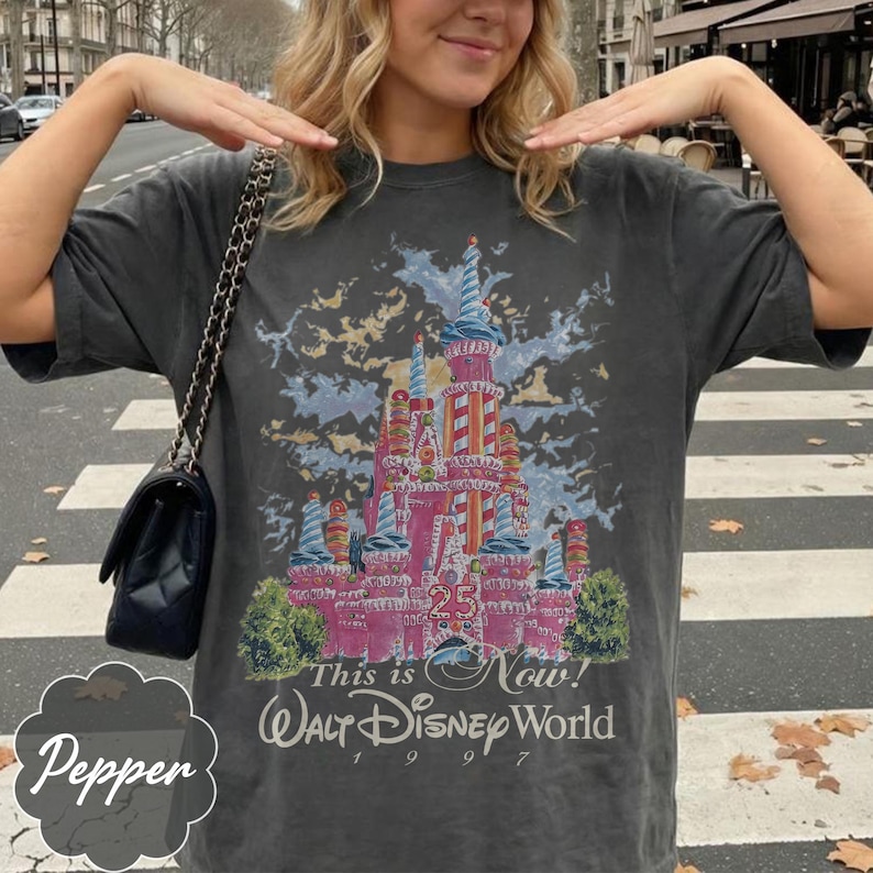 K&ouml;nnte beinhalten: Ein anthrazitfarbenes Kurzarm-T-Shirt mit einer Grafik eines rosafarbenen Schlosses mit bonbonartigen Akzenten. Der Text lautet "25", "This is Now!" und "Walt Disney World 1997". Eine schwarze gesteppte Handtasche mit silberner Kette h&auml;ngt &uuml;ber der Schulter.