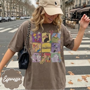 May include: A brown short-sleeve t-shirt featuring a grid of colorful cartoon characters from The Emperor's New Groove, including Kuzco, Pacha, Yzma, and Kronk, with the movie title text in the center, paired with a black quilted shoulder bag.