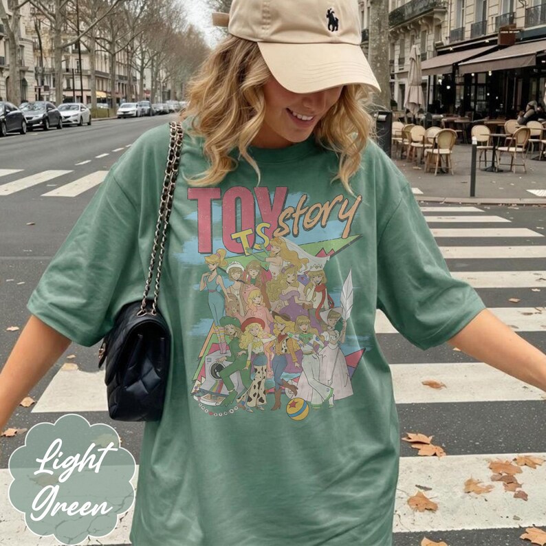 May include: A sage green short-sleeve t-shirt featuring a graphic print of various female characters from Toy Story with the text Toy Story in pink and yellow lettering. The shirt is paired with a black quilted shoulder bag with a chain strap.