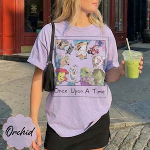 May include: A light purple t-shirt with a graphic of cartoon characters and the text "Once Upon A Time". The shirt is a crew neck style and has short sleeves. The shirt is worn with a black skirt.