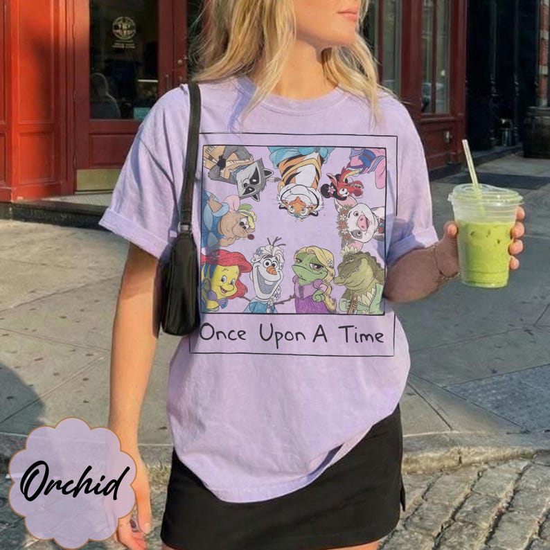 May include: A light purple t-shirt with a graphic of cartoon characters and the text "Once Upon A Time". The shirt is a crew neck style and has short sleeves. The shirt is worn with a black skirt.