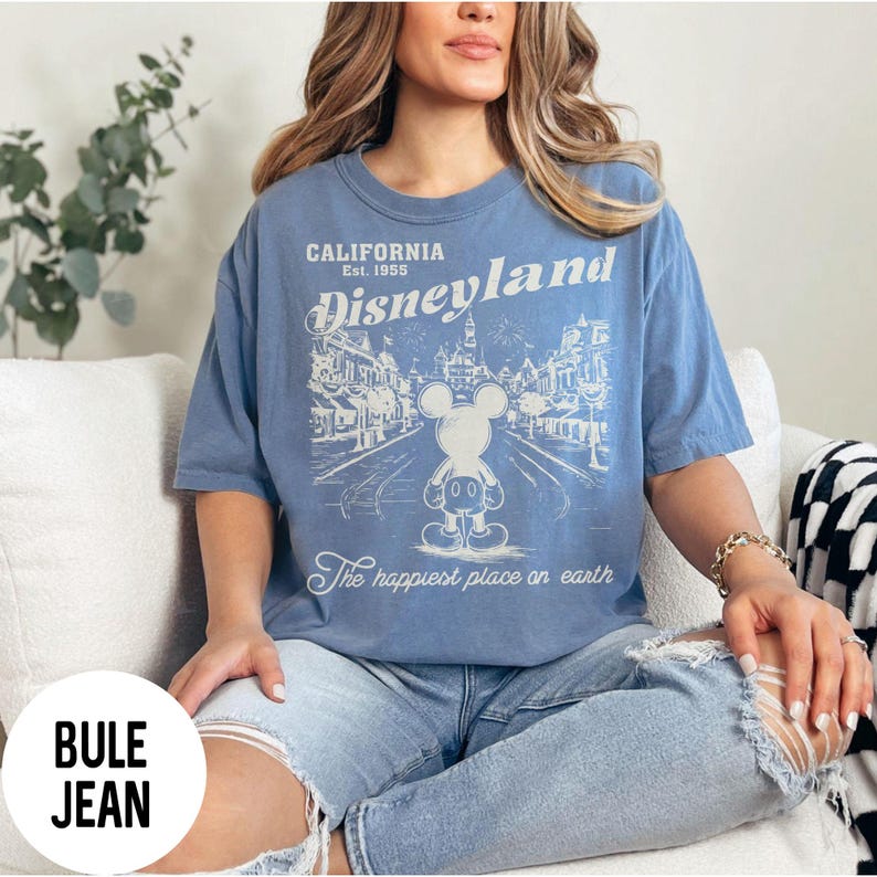 Może przedstawiać: Jasnoniebieska koszulka z grafiką Disneyland. Projekt zawiera tekst "California Est. 1955 Disneyland" oraz ilustrację Myszki Miki. Fraza "The happiest place on earth" jest r&oacute;wnież zawarta.