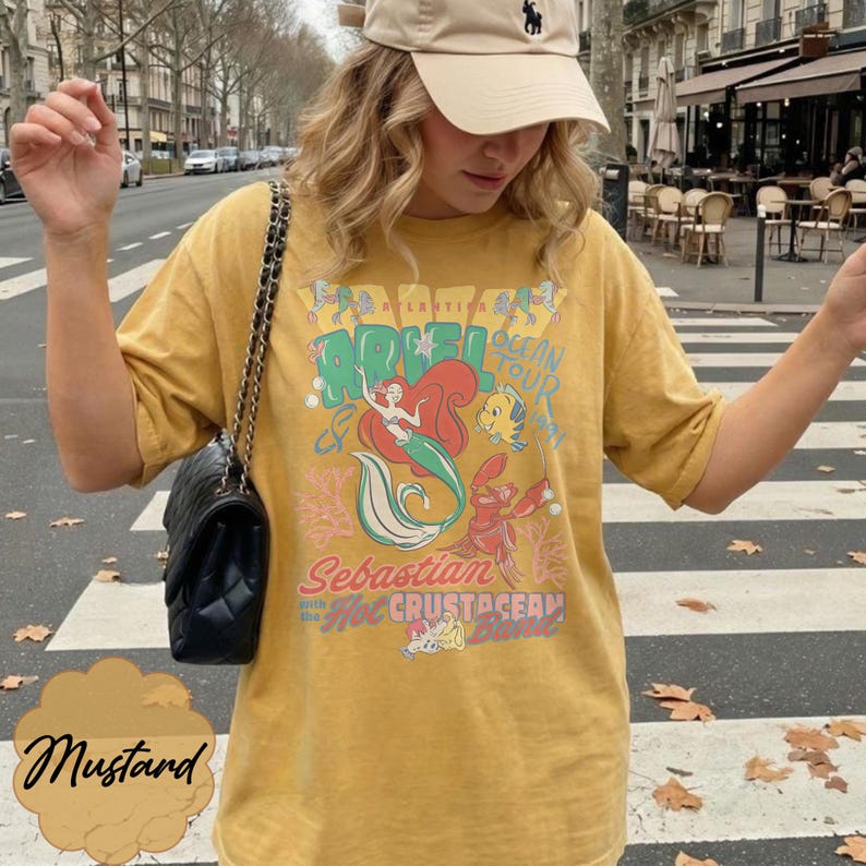 May include: Mustard-colored t-shirt featuring a graphic of Ariel from The Little Mermaid, with the words "Ariel Ocean Tour" and "Sebastian with the Hot Crustacean Band." The shirt has short sleeves and a relaxed fit.