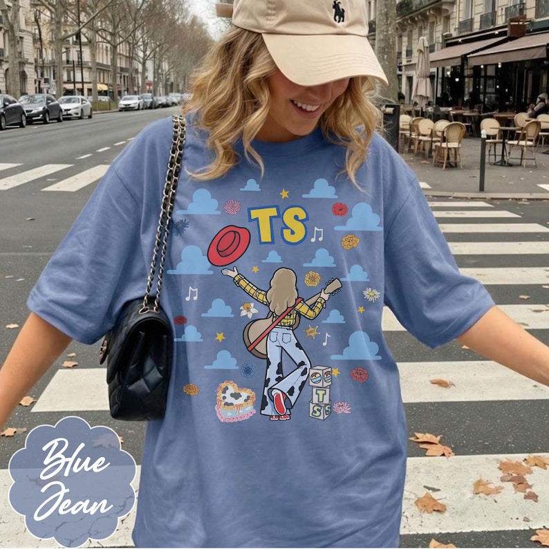 May include: A blue t-shirt featuring a graphic print of a person with a guitar, a red hat, and the text TS. The design includes blue clouds, musical notes, flowers, and cow-print patterns. The shirt has short sleeves and a loose, relaxed fit.
